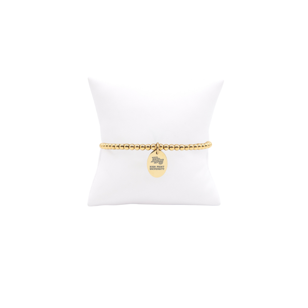 High Point University gold beaded bracelet with small oval charm with high point university text and hpu logo displayed on white pillow front view
