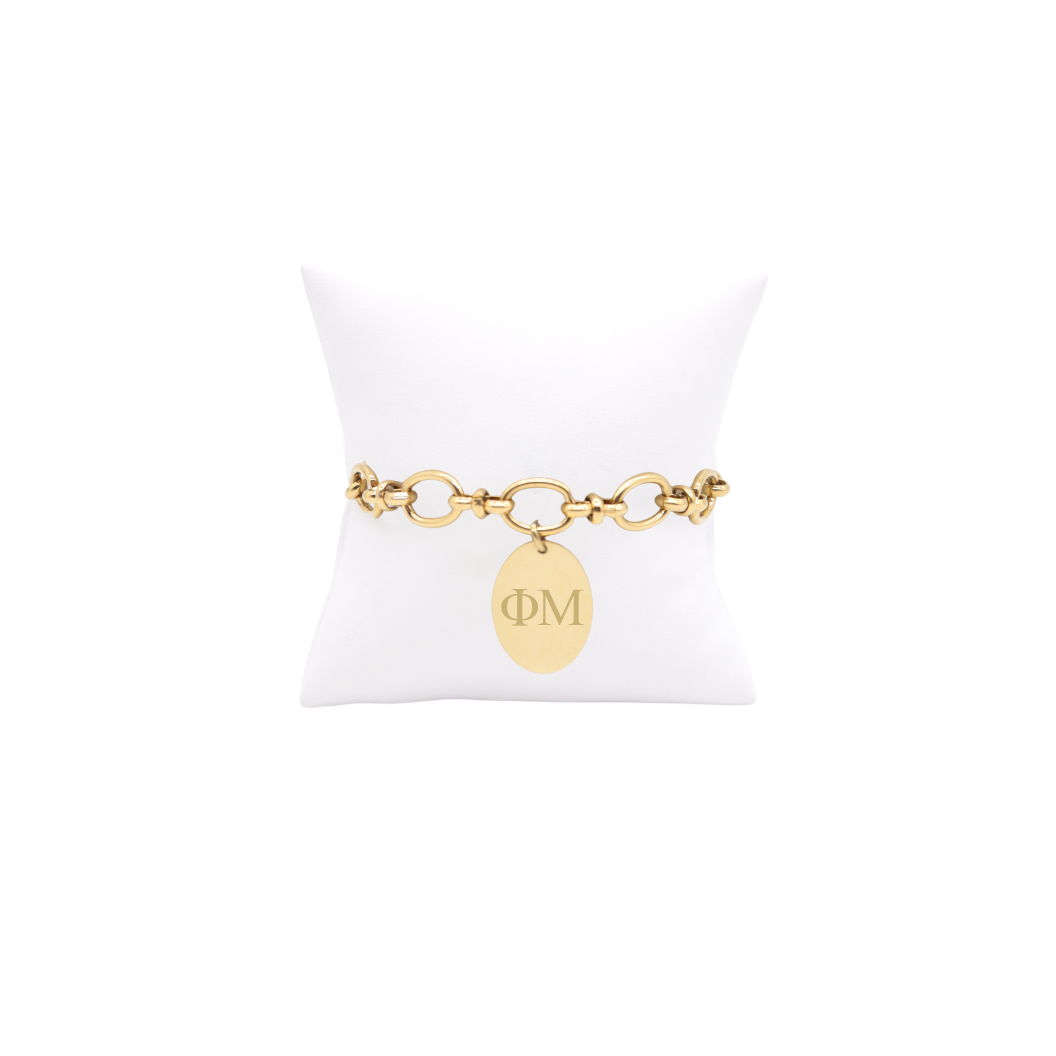 Phi Mu gold large link evermore bracelet with large oval charm with horizontal greek letters on white pillow front view
