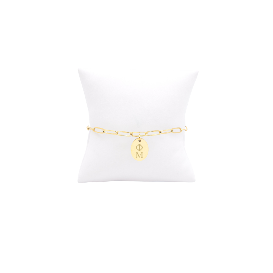 Phi Mu gold link storyline bracelet with small oval charm with vertical greek letters on white pillow front view
