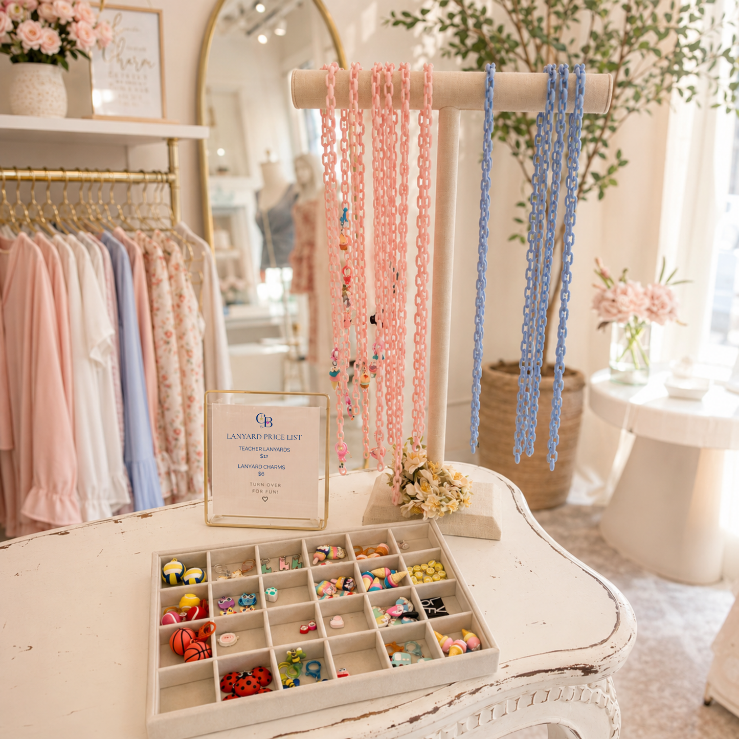 Teacher Lanyard Charm Bar display in a boutique featuring colorful pink and blue acrylic lanyard chains, a velvet tray of interchangeable teacher-themed charms, and a pricing sign on a white display table. Perfect for gift shops, boutiques, bookstores, and school spirit retailers.