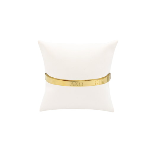 Alpha Chi Omega gold bangle bracelet with greek letters on white pillow front view.