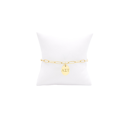 Alpha Sigma Tau gold link storyline bracelet with small oval charm displayed on white pillow front view with horizontal greek letters