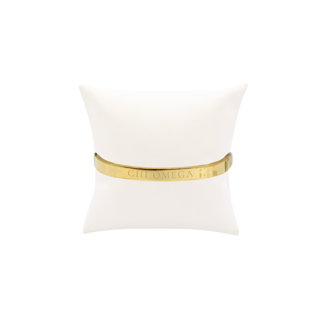Chi Omega gold bangle bracelet with full sorority name on white pillow front view.

