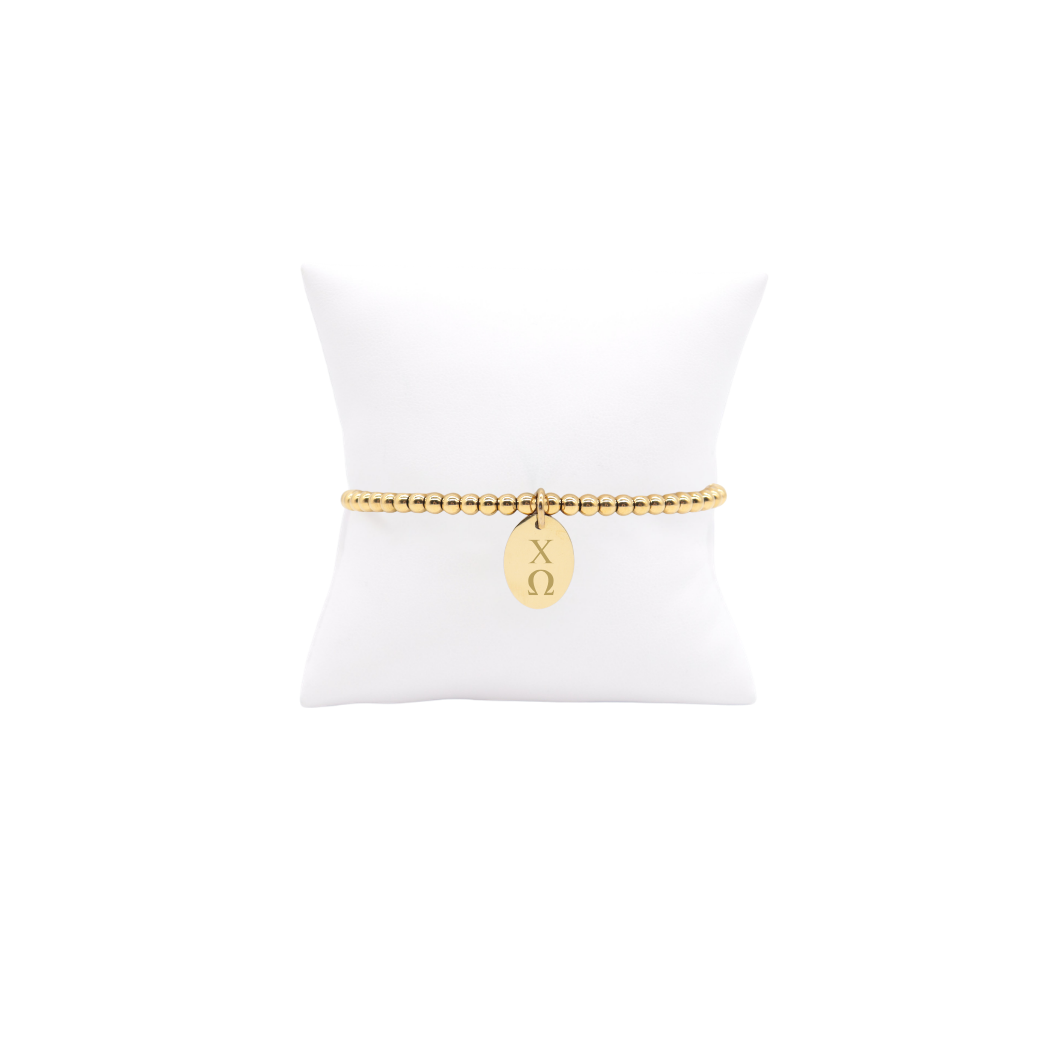 Chi Omega gold beaded bracelet with small oval charm with vertical greek letters displayed on white pillow front view