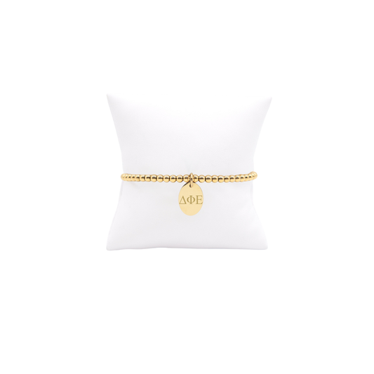 Delta Phi Epsilon gold beaded bracelet with small oval charm displayed on white pillow front view with horizontal greek letters