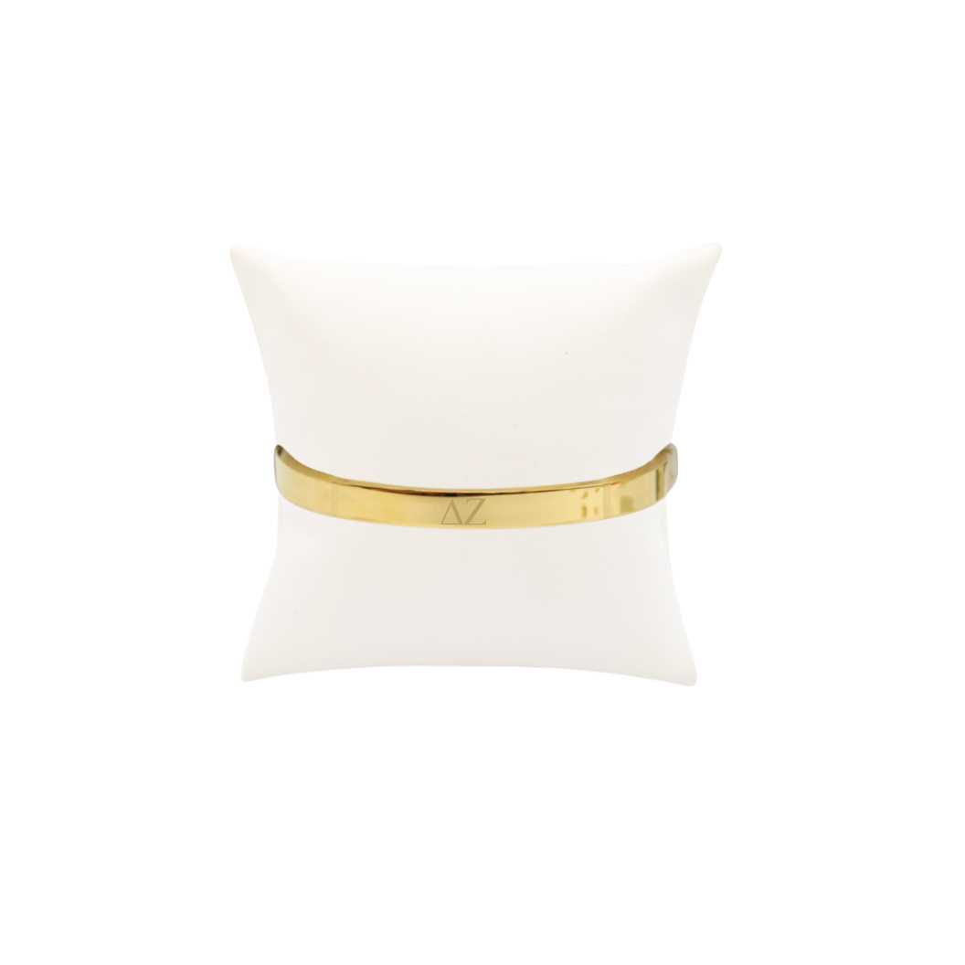 Delta Zeta gold bangle bracelet with greek letters on white pillow front view.
