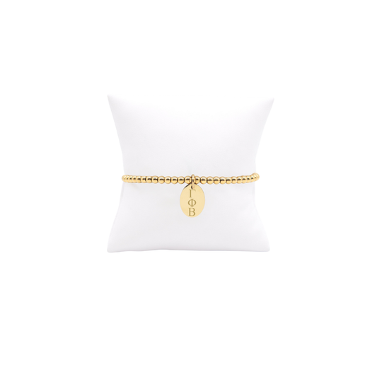 Gamma Phi Beta gold beaded bracelet with small oval charm displayed on white pillow front view with horizontal greek letters