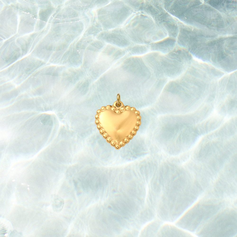 Gold heart charm with a polished center and beaded border on a shimmering water background.