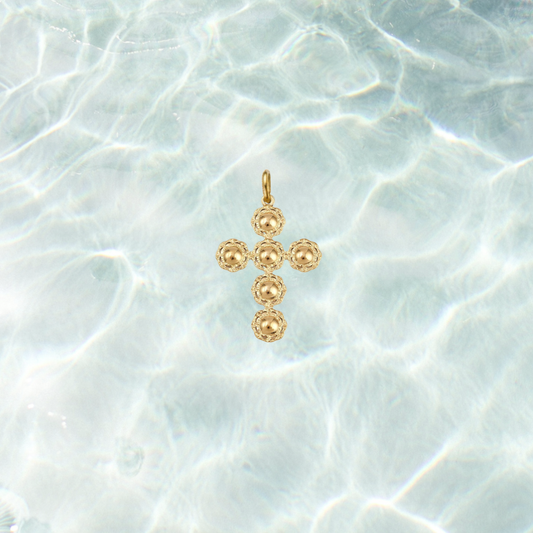 Gold filigree cross charm featuring a textured beaded cross design on a shimmering water background.