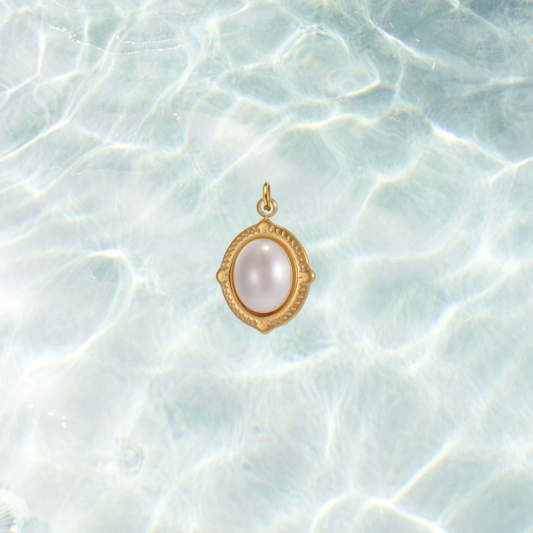 Gold oval charm featuring a luminous white pearl cabochon in a decorative bezel setting on a shimmering water background.