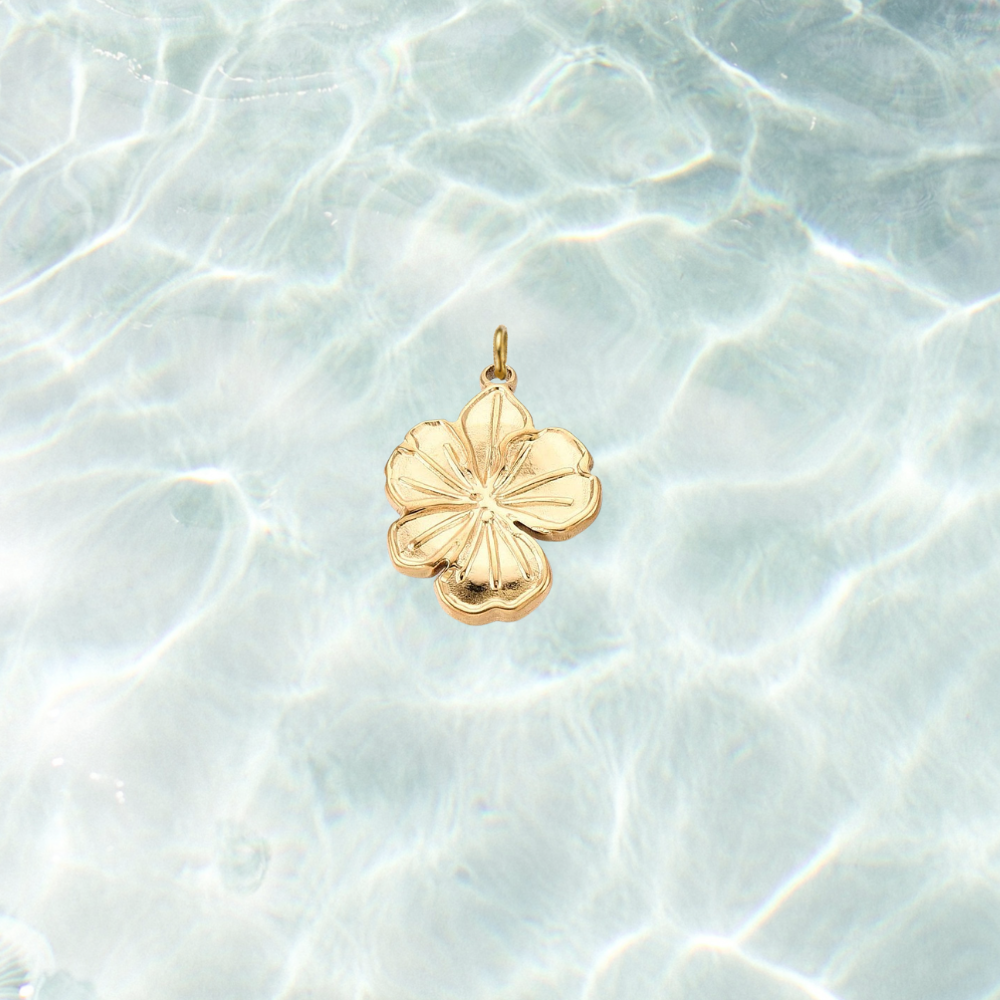 Gold hibiscus flower charm with engraved petal details and a polished finish on a shimmering water background.