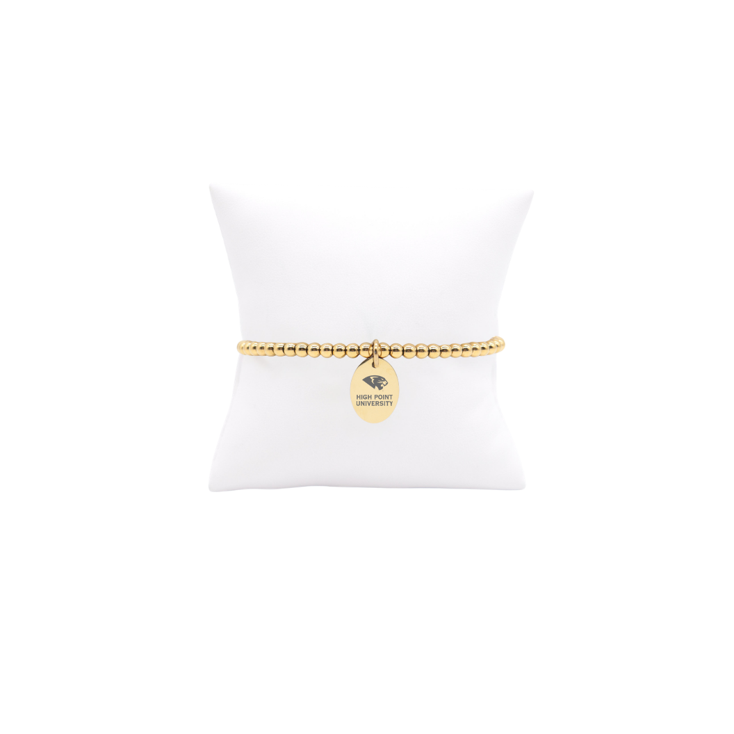 High Point University gold beaded bracelet with small oval charm with high point university text and panther logo displayed on white pillow front view

