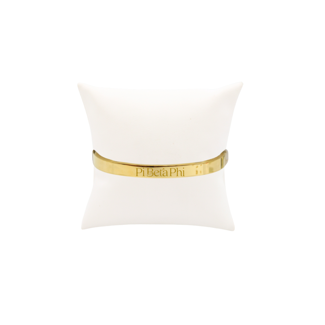 Pi Beta Phi gold bangle bracelet displayed on white pillow front view with new primary logo