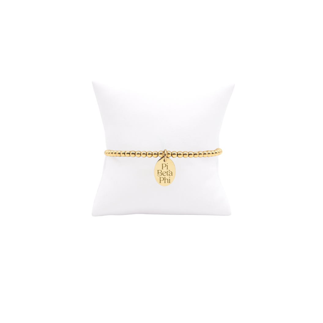 Pi Beta Phi gold beaded bracelet with small oval charm displayed on white pillow front view with full new stacked logo