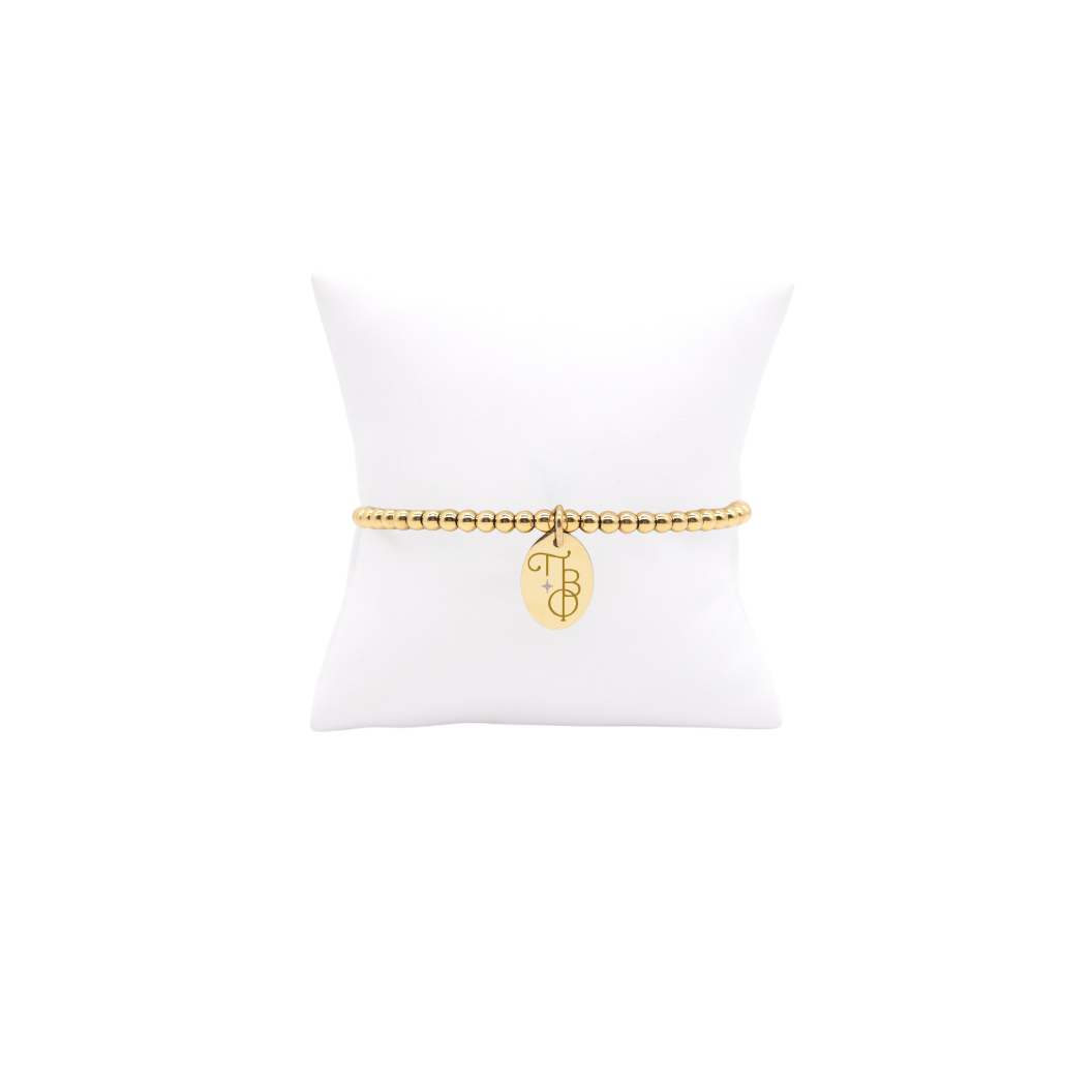 Pi Beta Phi gold beaded bracelet with small oval charm displayed on white pillow front view with new vertical/stacked greek letters