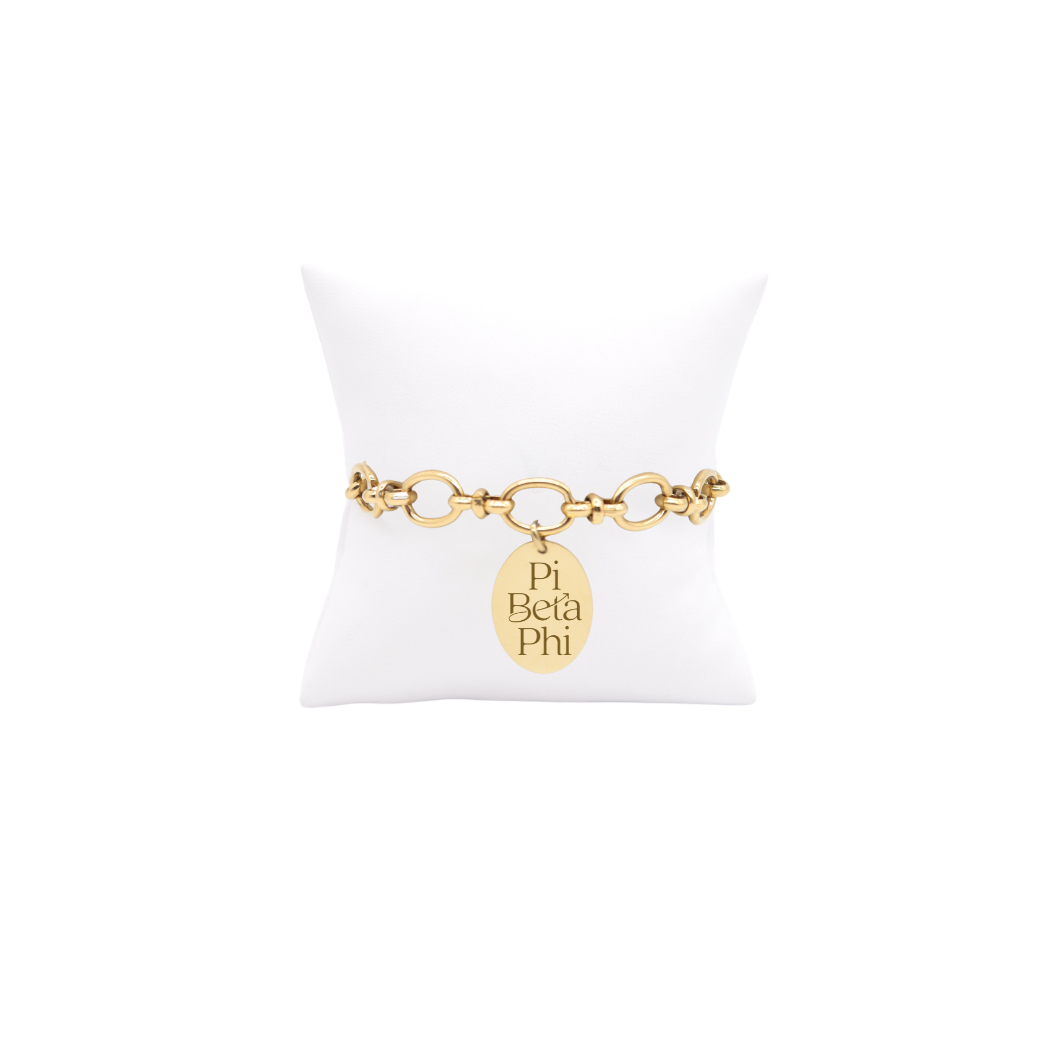 Pi Beta phi gold large link Evermore bracelet with large oval charm displayed on white pillow front view with new stacked logo