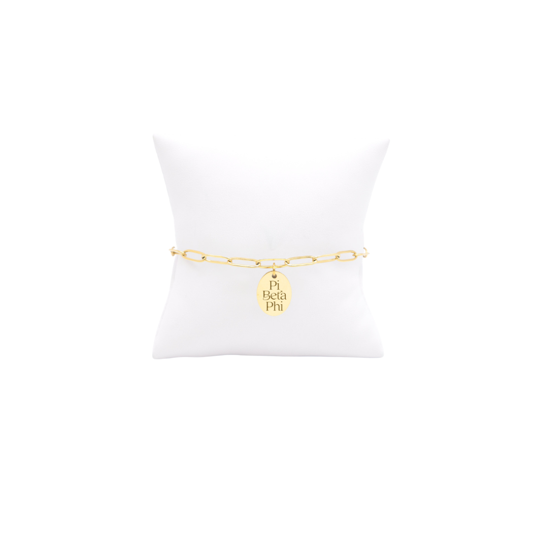 Pi Beta Phi gold link storyline bracelet with small oval charm displayed on white pillow front view with new stackd logo