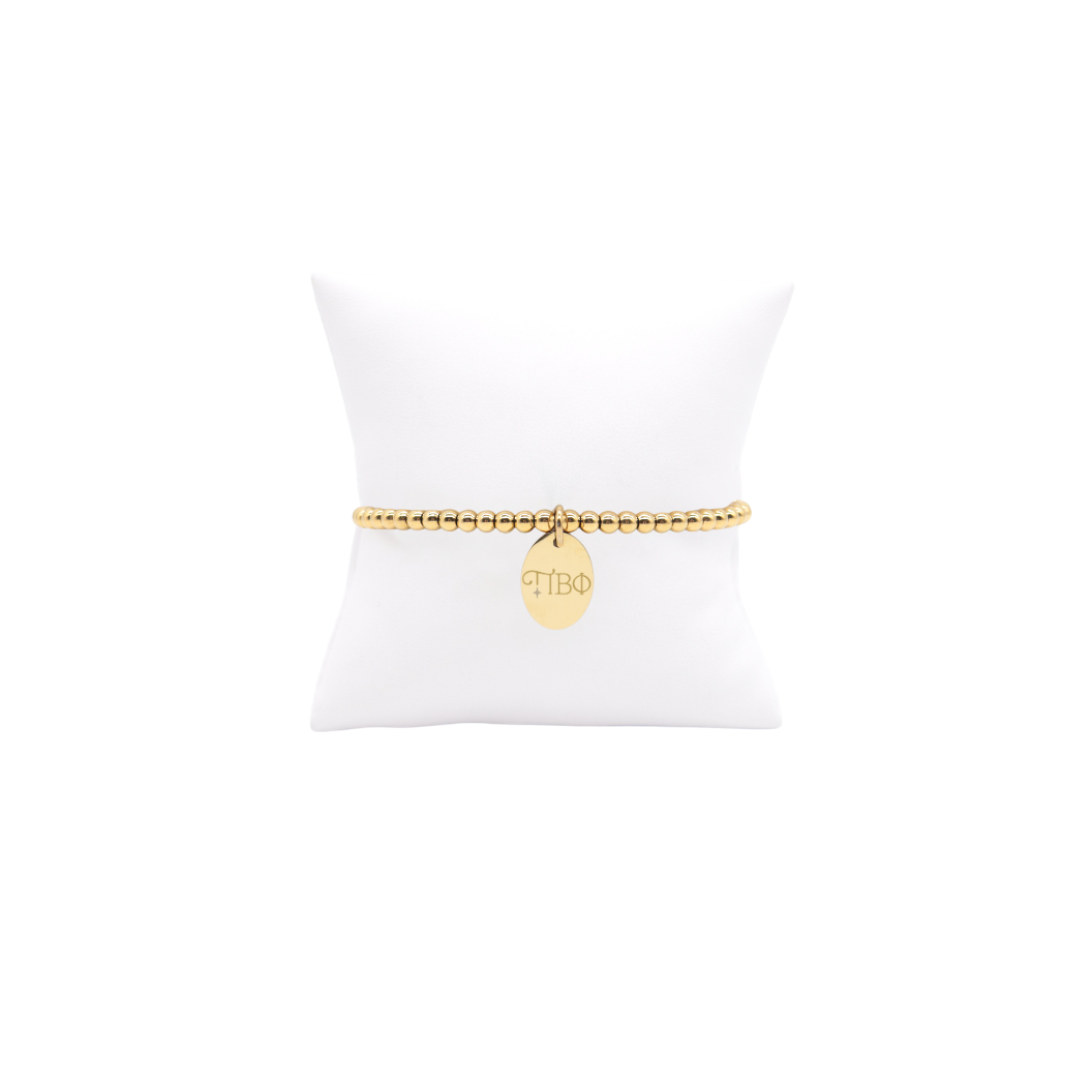 Pi Beta Phi gold beaded bracelet with small oval charm displayed on white pillow front view with new horizontal greek letters