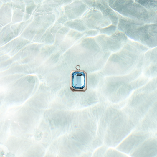 Silver rectangle charm featuring a faceted aqua blue gemstone on a shimmering water background.