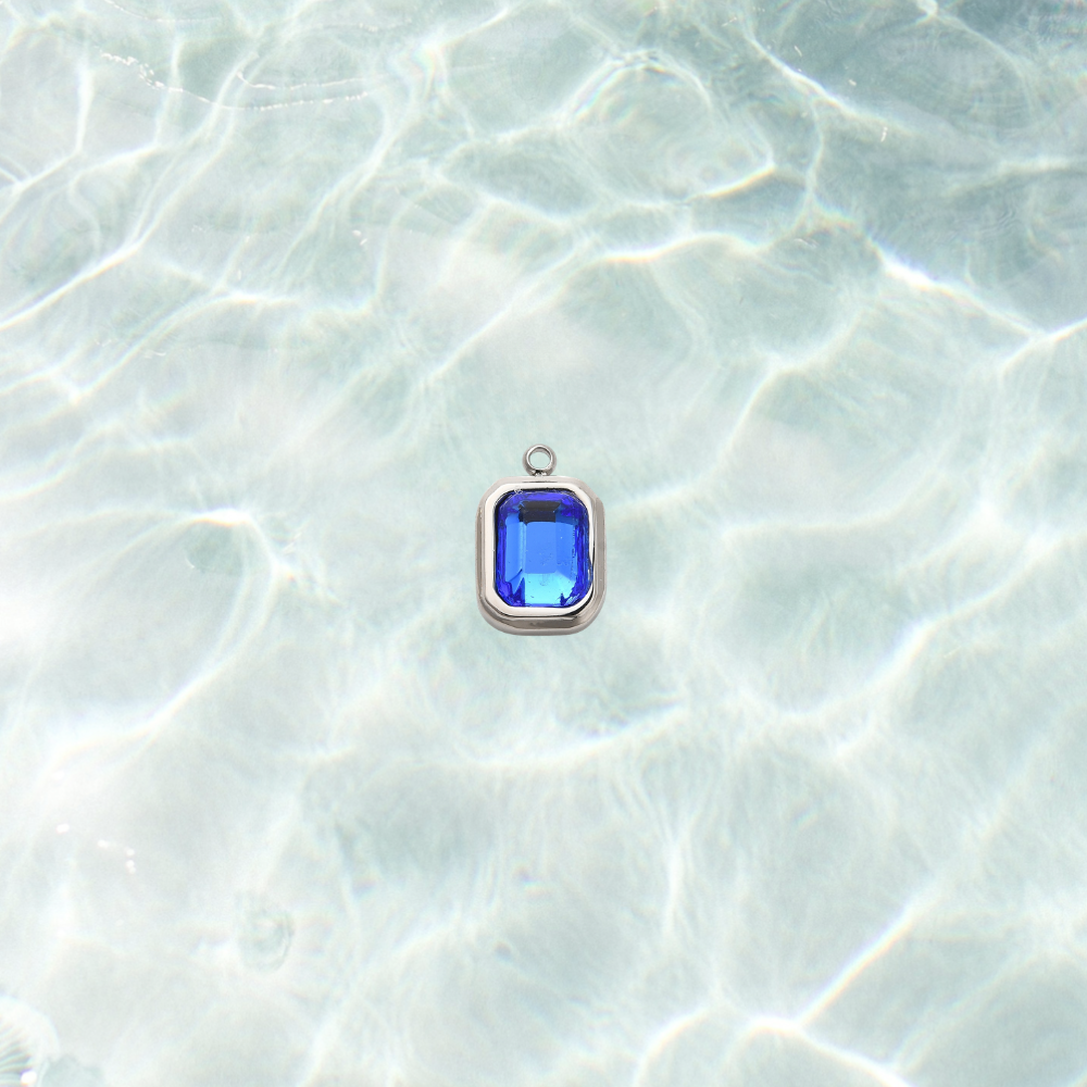 Silver rectangular charm featuring a faceted navy blue gemstone on a shimmering water background.