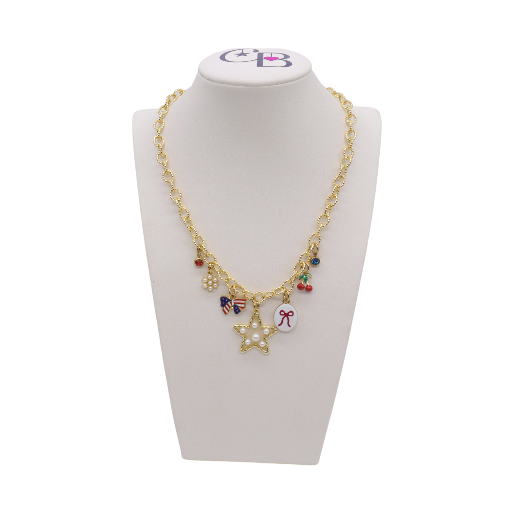 Gold charm necklace featuring a pearl star charm, patriotic bow charm, cherries, pearl cluster, bow illustration charm, and colorful crystal accents displayed on a white jewelry bust