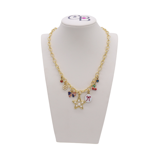 Gold charm necklace featuring a pearl star charm, patriotic bow charm, cherries, pearl cluster, bow illustration charm, and colorful crystal accents displayed on a white jewelry bust